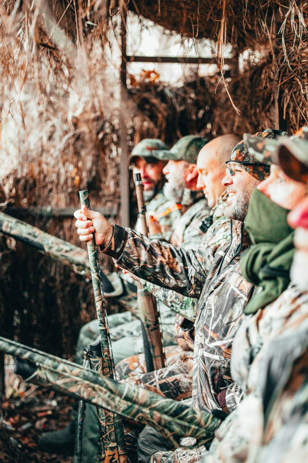 A group of hunters sit side by side inside a camouflaged blind made of brush and reeds. They wear matching camouflage clothing and gear, blending into their surroundings. Each holds a shotgun, ready for the hunt, while sharing focus and quiet anticipation. Warm light highlights their faces, adding a sense of camaraderie and patience to the moment.