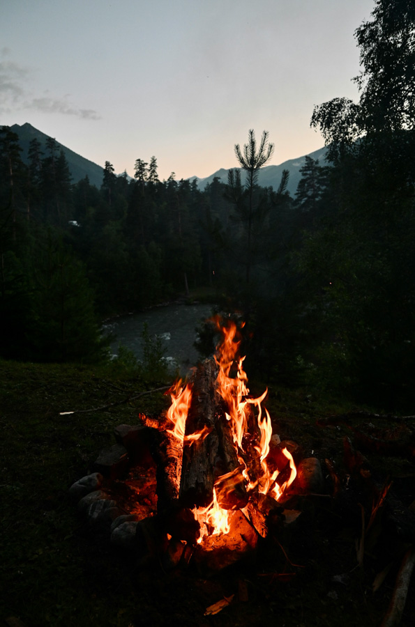 A glowing campfire burns brightly in the foreground, its flames curling around stacked logs and casting a warm light against the darkening evening. Behind it, a river winds through a dense forest of pine trees, with mountains silhouetted against the fading twilight sky. A serene wilderness scene full of warmth and solitude.