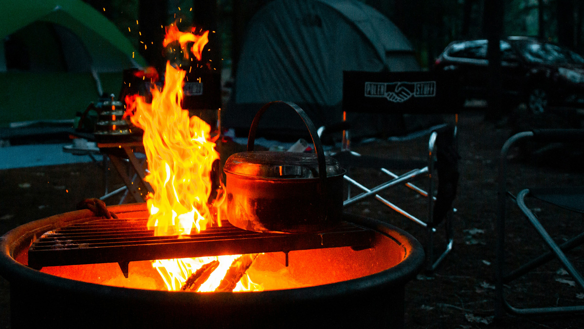 A lively campfire burns in a fire pit with a cast iron pot heating on a grill grate. The flames cast a warm glow against the dark forest backdrop, surrounded by camping gear, tents, folding chairs, and a parked car. The scene captures the rugged spirit of outdoor cooking and campfire gatherings.