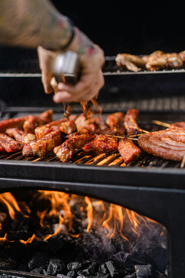 Close-up of meat grilling over open flames, with sausages and ribs on the grill grates while a person bastes the food with sauce.