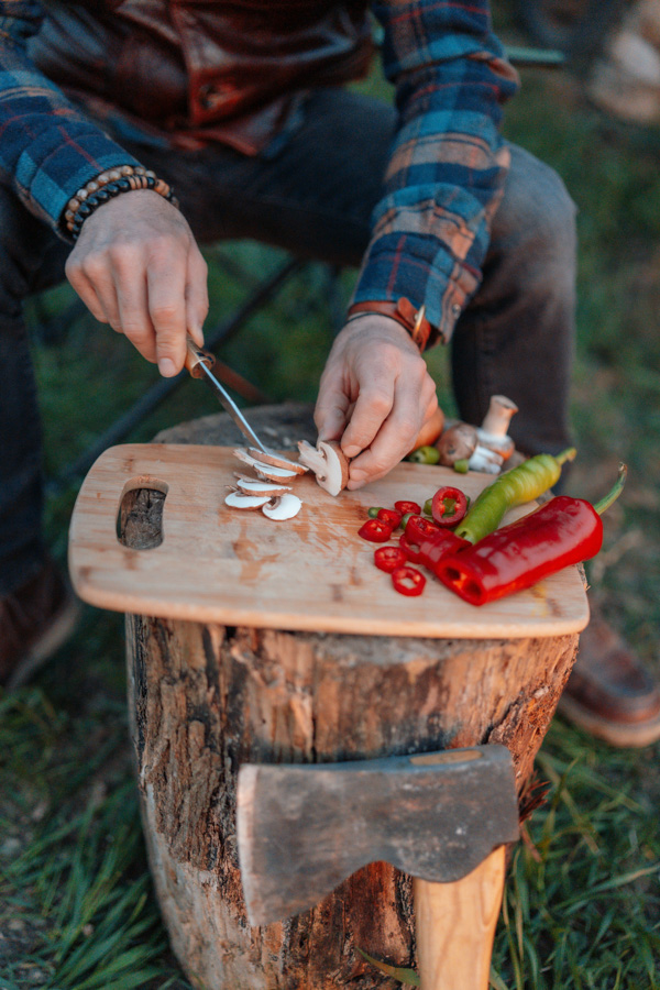 Person slicing mushrooms on a wooden cutting board placed on a tree stump, with red and green peppers nearby and an axe resting against the stump.