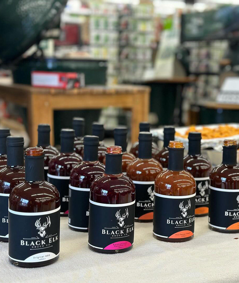 Display of Black Elk Sauce bottles on a table, with varieties including Original BBQ, Cranberry Habanero, and Hot Pepper Peach, each featuring a black label with a white elk head logo.