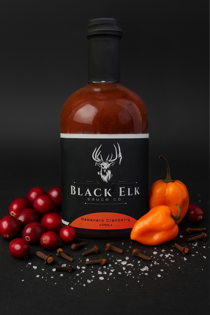 Bottle of Black Elk Sauce Co. Habanero Cranberry Sauce (375mL). Displayed with fresh cranberries, orange habanero peppers, cloves, and coarse sea salt against a dark backdrop. The black label with stag emblem and red accent conveys a bold, seasonal blend of sweet-tart cranberry and fiery habanero heat.