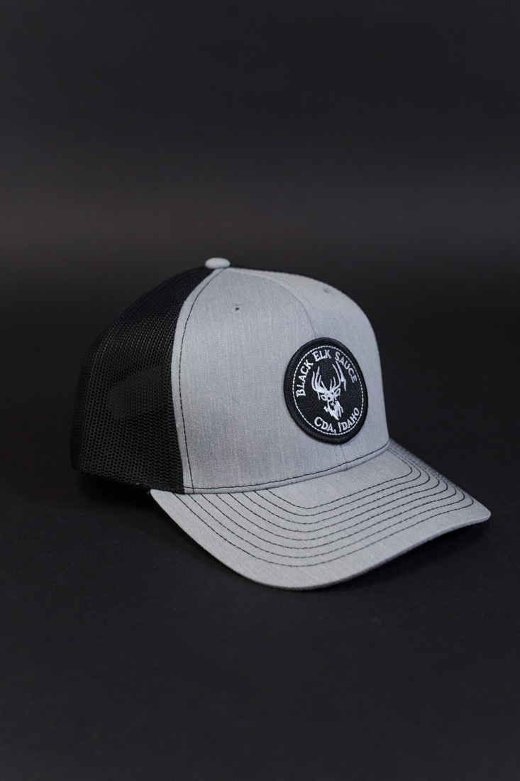Gray and black mesh trucker hat with a round Black Elk Sauce patch on the front, featuring a white elk head logo and text reading “Black Elk Sauce CDA, Idaho.”