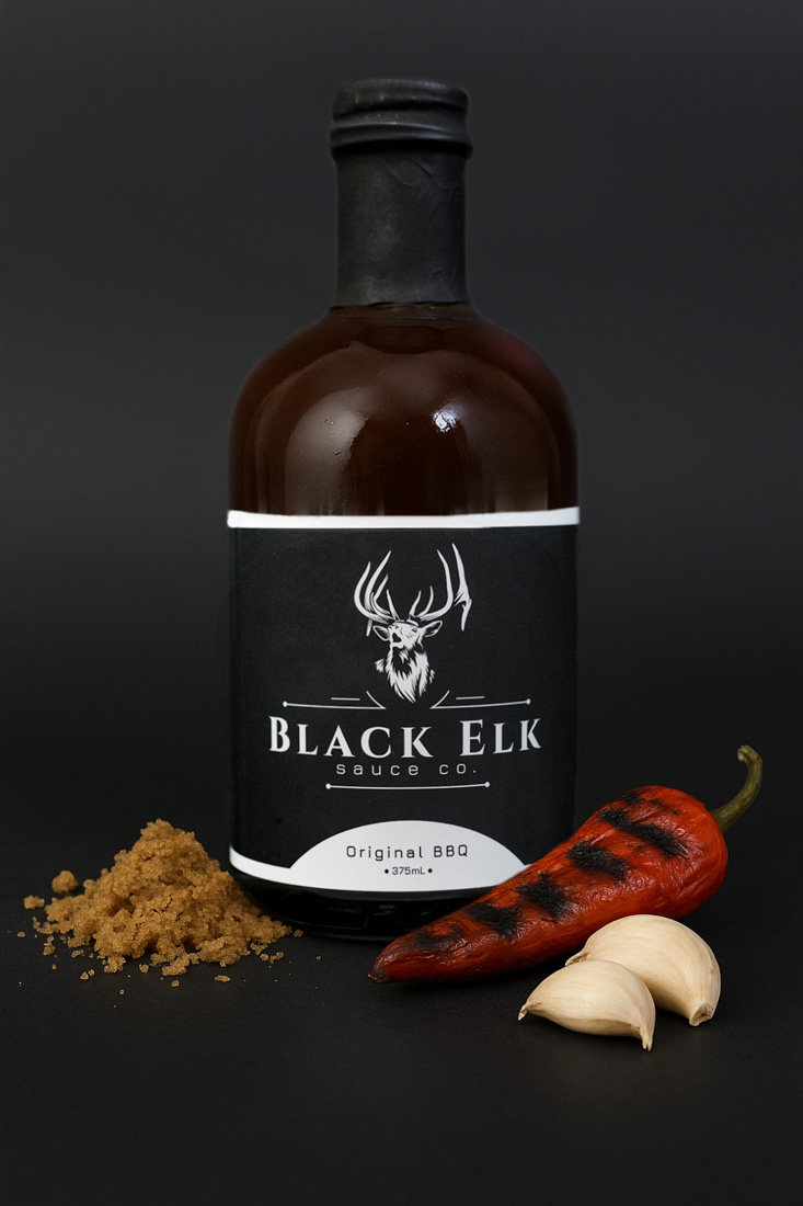 Bottle of Black Elk Sauce Co. Original BBQ Sauce (375mL). The label showcases the signature elk head logo in bold white on black. Styled with a rustic spread of brown sugar, char-grilled red pepper, and garlic cloves, the scene highlights the sauce’s balance of sweet, smoky, and savory BBQ flavors.