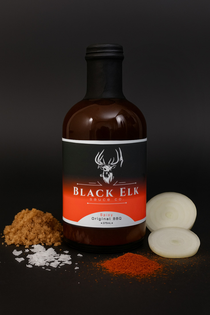 Bottle of Black Elk Sauce Co. Spicy Original BBQ Sauce (375mL). Brown glass bottle with a black-to-red gradient label featuring the white elk head logo. Displayed with raw ingredients — brown sugar, sea salt, paprika, and sliced onion — emphasizing the bold, flavorful blend of spices used in the sauce.