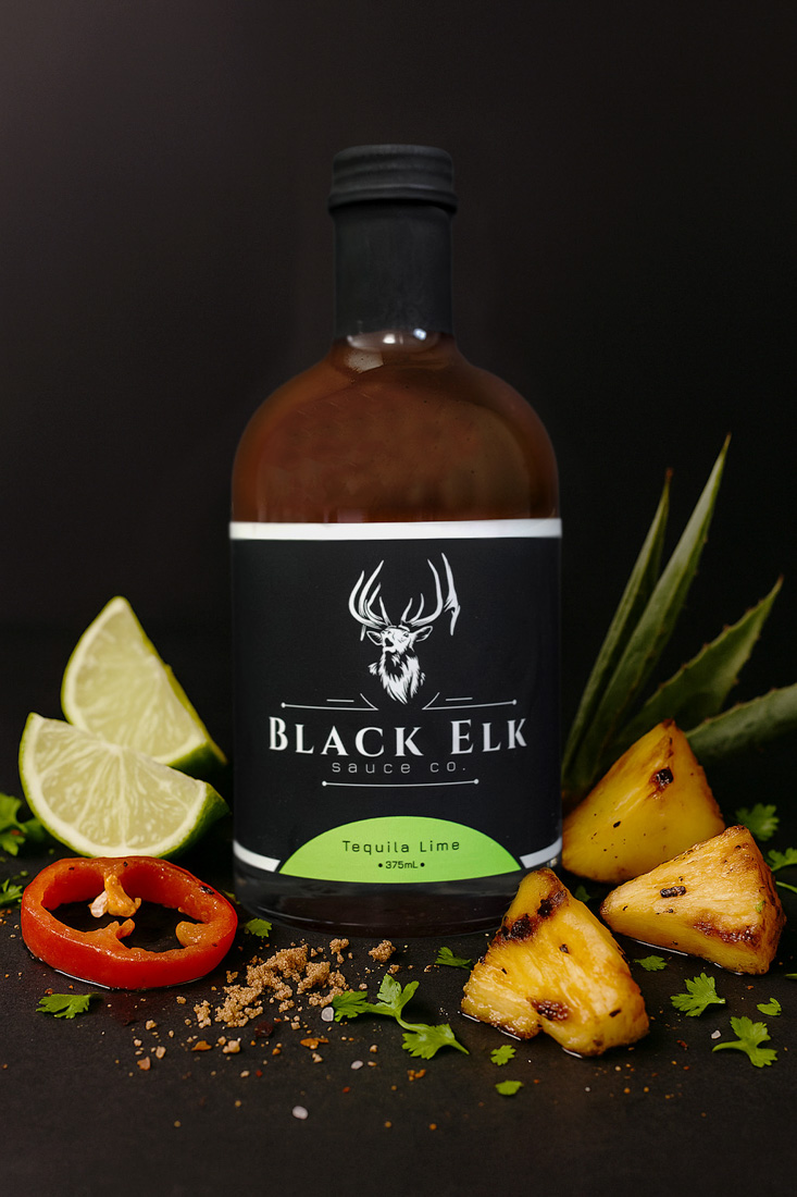 Bottle of Black Elk Sauce Co. Tequila Lime BBQ Sauce (375mL). Brown glass bottle with a black label featuring a white elk head logo. The flavor "Tequila Lime" is highlighted in bright green. Styled with lime wedges, grilled pineapple chunks, red chili slices, cilantro, and brown sugar on a dark background for a fresh, tropical presentation.