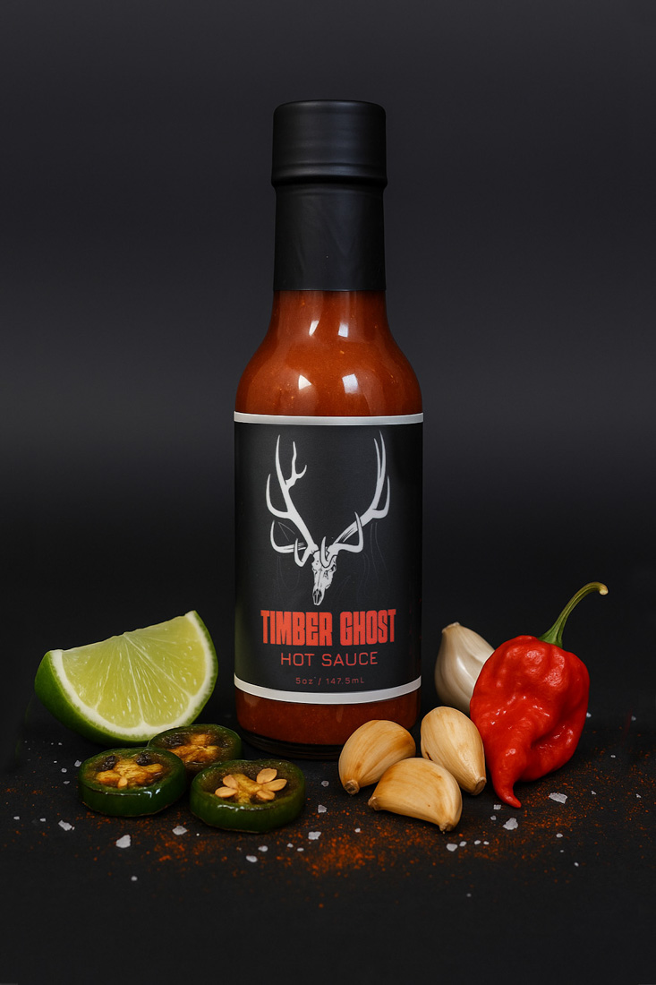 Bottle of Timber Ghost Hot Sauce (5oz / 147.5mL) with black label featuring a white deer skull logo and bold red text. The bottle is surrounded by fresh ingredients including lime, jalapeño slices, garlic cloves, a red chili pepper, and sprinkled salt and chili powder, set against a dark background.