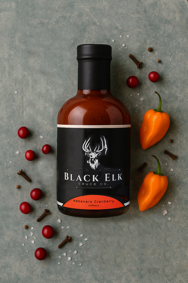 A bottle of Black Elk Habanero Cranberry sauce rests on a textured surface, surrounded by fresh cranberries, bright orange habanero peppers, whole cloves, and scattered coarse salt. The arrangement highlights the sauce’s balance of fiery heat and tart, fruity depth.