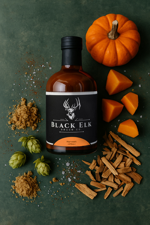 A bottle of Black Elk Harvest sauce is displayed on a dark green surface, surrounded by seasonal ingredients. A small pumpkin with bright orange chunks, wood chips, hops, brown sugar, and coarse salt frame the scene, highlighting the sauce’s autumn-inspired, earthy, and slightly sweet flavor profile.