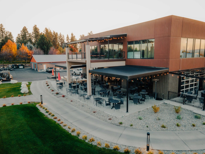 Modern two-story restaurant with large windows and outdoor patio seating on both levels, surrounded by landscaped walkways and green lawn.