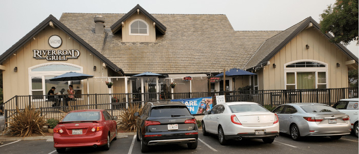 Riverroad Grill restaurant exterior with beige siding, peaked roof, patio seating under blue umbrellas, and several cars parked in front.