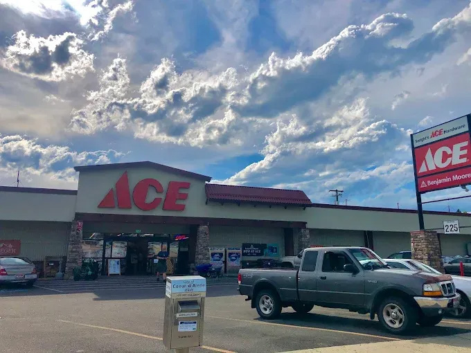Ace Hardware store with a red ACE sign above the entrance, a pickup truck parked in front, and a roadside sign showing Ace and Benjamin Moore branding under a cloudy blue sky.