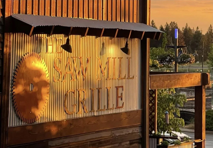 Sign for The Saw Mill Grille on a corrugated metal wall with a large circular saw blade design, lit by warm sunset light