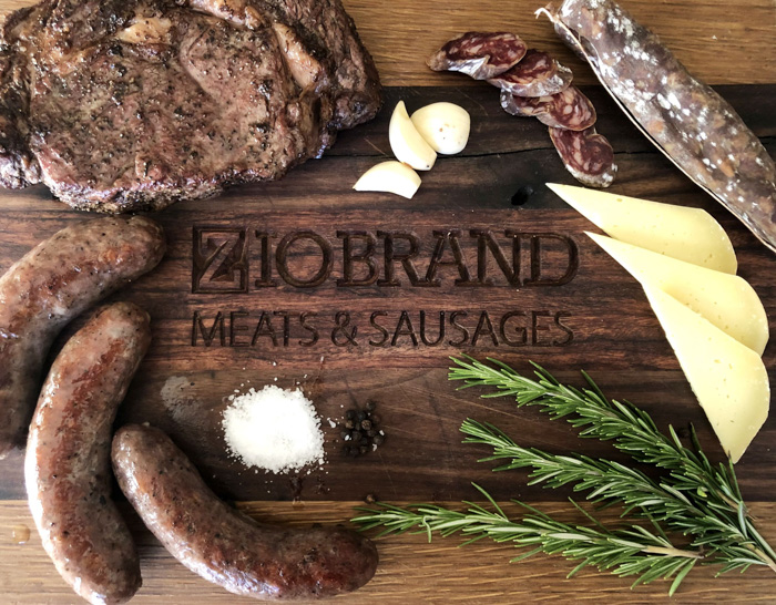 Wooden cutting board engraved with 'Ziobrand Meats & Sausages,' surrounded by grilled steak, sausages, sliced salami, cheese wedges, garlic cloves, rosemary sprigs, black pepper, and coarse salt