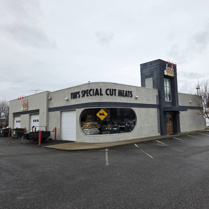 Exterior of Tim’s Special Cut Meats, a gray building with large front windows, garage doors, and bold black signage above the entrance